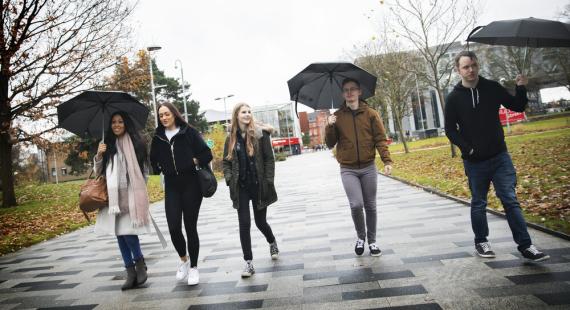 Group of 5 students walking along the University broadwalk in the rain, holding umbrellas.