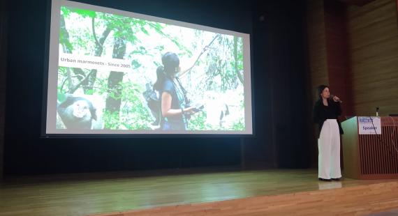 University Fellow, Marina Duarte, stood on stage holding a microphone with a screen behind her showing a picture of a forest