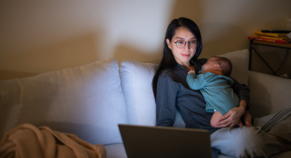 A mum sat working on her laptop while holding her child who is sleeping