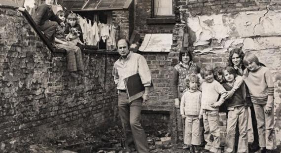 photograph of a group of children and an adult man standing in a rundown urban courtyard with brick buildings and broken windows in the background, suggesting poor housing conditions.