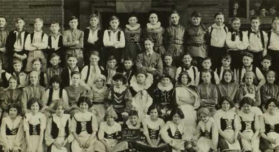 Black-and-white group photograph of approximately 60 schoolchildren and several adults posed in four rows outside a brick building. Many of the children wear coordinated costumes with dark vests or bodices over light shirts, suggesting a school performance or class production.