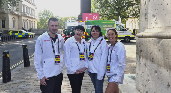 A group of four podiatry students and staff smile at the camera. They are stood on the street in front of a number of support vehicles and wear official London Marathon branded jackets and lanyards.