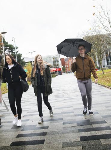 Group of 5 students walking along the University broadwalk in the rain, holding umbrellas.
