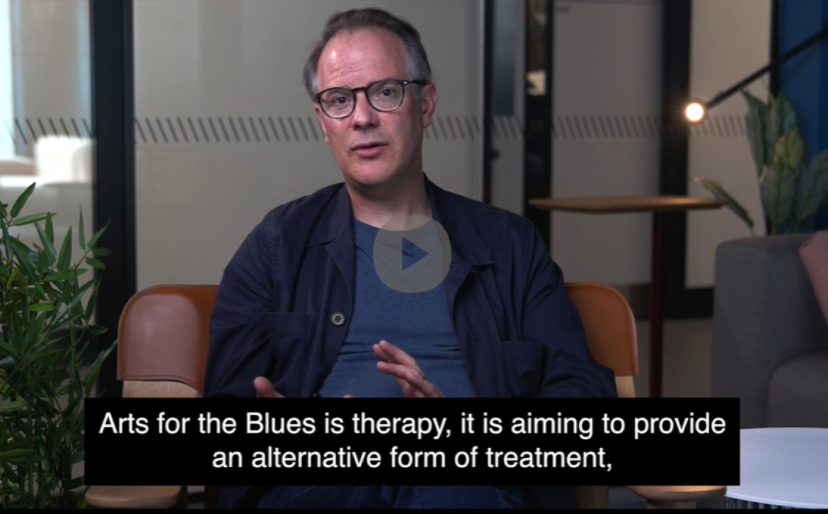 Scott Thurston (middle-aged, white man, with glasses) sitting in a mustard coloured chair, plant on the left hand side, settee to the right